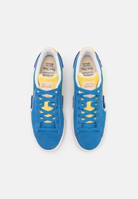 Modré semišové tenisky s žlutými akcenty a tkaničkami. Mají bílou mezipodešev a texturovaný design. Logo Puma je umístěno na jazyku.