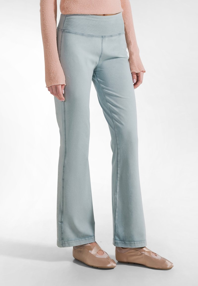 Mujer con pantalones acampanados azul claro, una blusa de manga larga color melocotón con agujeros para los pulgares y bailarinas beige con tiras cruzadas.