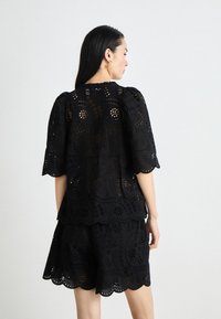 Top in cotone nero con disegno a pizzo ritagliato, maniche puffate e orlo scallop, abbinato a pantaloni corti in tessuto e dettaglio simili.