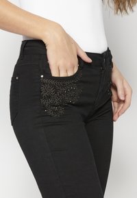 Black jeans feature embroidered floral designs in gold tones on the front pocket. The material appears stretchy, with silver hardware accents.