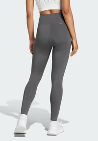 Rear view of a person wearing high-waisted gray leggings, white sneakers, and a white cropped top, standing on a light background.