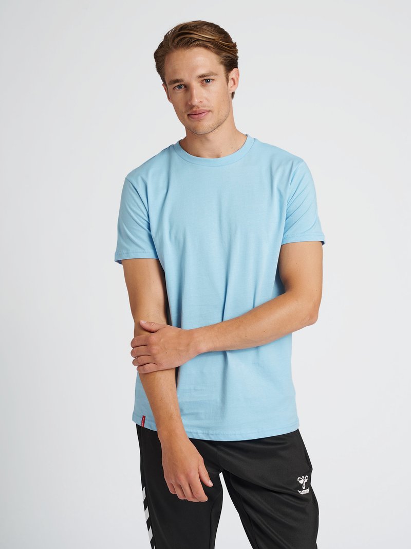 Jonge man in lichtblauw T-shirt met korte mouwen en zwarte sportbroek, staand tegen een effen witte achtergrond.