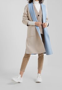 Beige lange jas met een lichtblauwe kraag, gecombineerd met een beige sweater vest, bruine corduroy broeken en witte sneakers. Glad materiaal en een getailleerde pasvorm.
