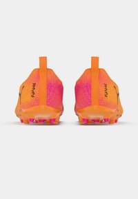 Pāris oranžām un rozā futbolam paredzētām apavām ar melnu Puma logo, skatīts no aizmugures uz balta fona.