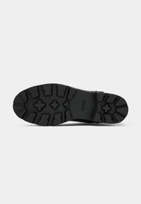 Suela de zapato de goma negra con un patrón texturizado que presenta surcos profundos, formas moldeadas y el nombre de la marca "GUESS" visible.