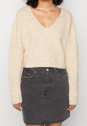 Personne portant un pull en maille beige col en V et une jupe en denim gris foncé taille haute sur un fond uni.
