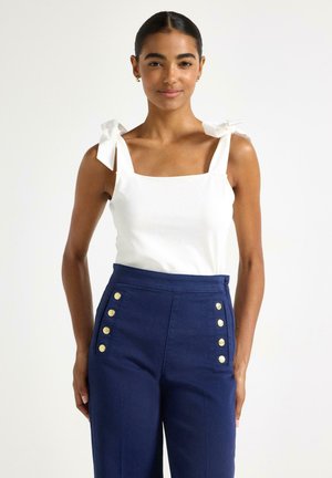 Giovane donna che indossa un top bianco senza maniche con spalline a fiocco e pantaloni blu navy a vita alta con bottoni dorati, in piedi su sfondo bianco.