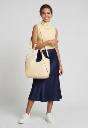 Smiling woman wearing yellow sleeveless turtleneck, navy midi skirt, white sneakers, carrying large beige shoulder bag against plain background.
