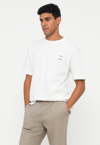 White short-sleeve cotton t-shirt featuring a subtle brand logo in dark blue on the left side, paired with light beige pants.