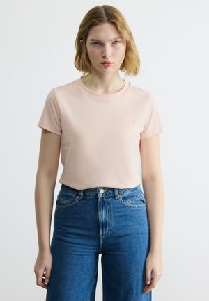 Νεαρή γυναίκα με ξανθά μαλλιά μέχρι τους ώμους, φορώντας ανοιχτό ροζ μπλουζάκι με κοντά μανίκια, μπλουζωμένο μέσα σε ψηλόμεσο μπλε τζιν, μπροστά σε απλό φόντο.