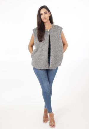 Grå faux-fur vest med ærmeløs design, lige snit og åben front. Båret over en sort top, parret med blå jeans og klare sandaler.