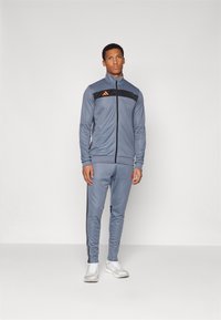 Grijze sportieve trainingsoutfit met een ritssluiting jasje met oranje accenten en zwarte panelen, gecombineerd met bijpassende broek en witte sneakers.