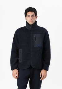 Jachetă de fleece bleumarin cu o suprafață texturată, guler înalt, față cu fermoar și trei buzunare. Dispune de un logo pe buzunarul din față.