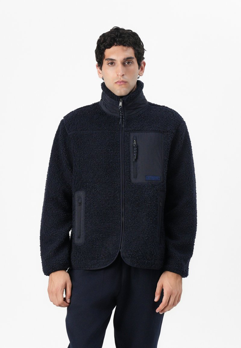 Jachetă de fleece bleumarin cu o suprafață texturată, guler înalt, față cu fermoar și trei buzunare. Dispune de un logo pe buzunarul din față.