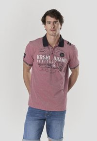 Punane polo-särk tume kraega, millel on trükitud tekst ja ümmargune logo. Kombineeritud hele siniste teksapükstega, kujutatud ühevärvilise tausta ees.