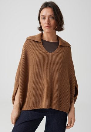 Femme aux cheveux bruns courts portant un poncho en maille marron ample et côtelé par-dessus un haut marron foncé et un jean foncé, sur un fond uni.