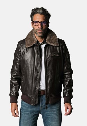 Homme portant une veste en cuir marron foncé avec un grand col en fourrure, une chemise blanche, un jean bleu et des lunettes, debout devant un fond uni.