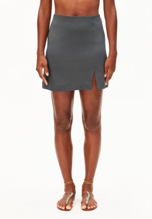 Femme portant une mini-jupe gris foncé avec une fente latérale et des sandales marron à brides à la cheville, debout devant un fond blanc.