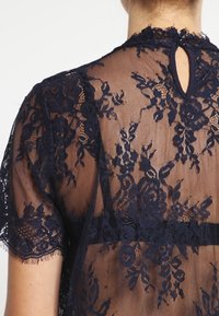 Blusa in pizzo blu navy con motivi floreali, tessuto trasparente, maniche corte e dettaglio a chiave sulla schiena chiuso da un bottone.