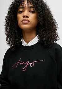 Felpa nera con scollo a girocollo, caratterizzata da una scritta rosa lucida "Hugo" sulla parte anteriore. Tessuto testurizzato e maniche raglan.