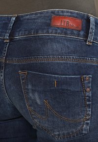 Mörkblå denimjeans med en läderetikett som har "LTB", kontrasterande gul sömnad och en bakficka med en dekorativ söm.