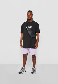 Camiseta negra oversize con un gráfico de un pájaro blanco, combinada con shorts morados claros y zapatillas deportivas negras con detalles grises. Bolso bandolera en negro.