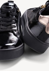 Scarpe in vernice nera con suola spessa, dotate di lacci rotondi, dettagli dorati e un elemento metallico a contrasto su un lato.