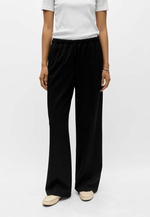 Personne portant un pantalon noir ample avec une taille élastique et des chaussures blanches de style mocassin, debout devant un fond uni.