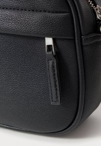 Μαύρη τσάντα από δέρμα με υφή, με φερμουάρ κλεισίματος, που διαθέτει ασημί μεταλλική λεπτομέρεια και μικρό δερμάτινο τράβηγμα. Λείο και συμπαγές σχέδιο.