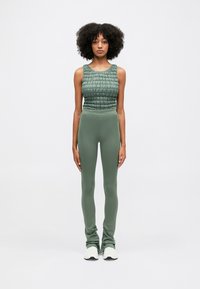 Grøn cropped tanktop med tekstureret stof, kombineret med tætsiddende grønne leggings. Modellen bærer hvide sportssko. Ensfarvet, glat tekstur.