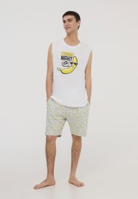 Canotta bianca senza maniche con scritta "TOUGH NIGHT" e grafica di una banana. Pantaloncini gialli e grigi con motivi a banana. Modello a piedi nudi.