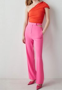 Frau in einem einseitigen, orangefarbenen Oberteil, kombiniert mit hochtaillierten, rosa Hosen und passenden, spitzen, rosa Schuhen, die vor einer schlichten Wand steht.