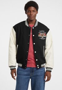 Mustvalge varsity jakk, mis on nööpidega, triibuliste varrukate otstega ja tikandiga rinnalogoga. Kanti punase särgi ja siniste teksade peal.
