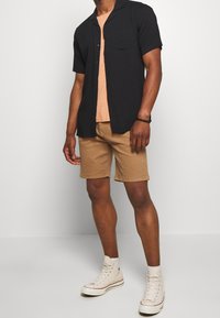 Chemise noire à manches courtes, t-shirt beige en dessous, short beige et baskets blanches, présentant une tenue décontractée avec des couleurs contrastées.
