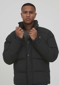 Black puffer jacket with a high collar, snap fasteners, and quilted design. Model is holding the collar, revealing a gray inner layer.