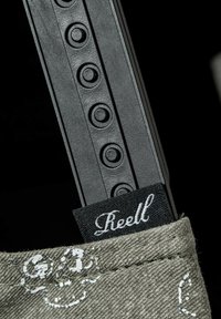 Black plastic item with circular impressions resting in a gray fabric pocket featuring a white skull pattern and a black "Reell" label.