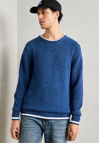 Blå strikket genser med ribbestrikkede mansjetter og kant, med en hvit aksentstripe. Kombinert med lyseblå jeans og en svart caps.