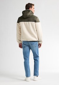 Beige fleece jas met een olijfgroen bovenstuk en capuchon. Lichtblauwe jeans en witte sneakers maken de outfit compleet.