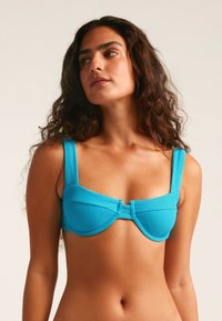 Top de bikini de color turquesa con costillas, tirantes gruesos, soporte de varilla y detalle de lazo en el centro. Textura suave y diseño estructurado.
