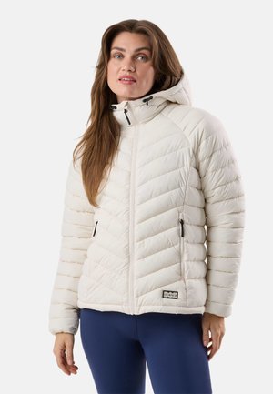 Woman wearing a white padded jacket with a hood and zippered pockets, paired with navy blue leggings, standing against a plain background.