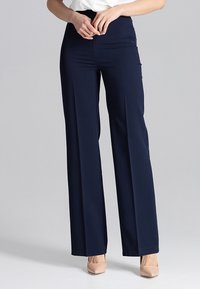 Vrouw in hoge taille marineblauwe wijde broek met beige puntige hakken, met de handen op de taille gekruist, staand op een effen achtergrond.