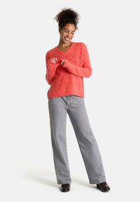 Korallenfarbener Strickpullover mit V-Ausschnitt und strukturiertem Muster, kombiniert mit hellgrauen weit geschnittenen Hosen und dunklen Schuhen.