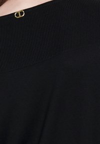 Černý pletený svetr s vroubkovaným výstřihem a malým zlatým detailem na zádech. Hladká textura a uvolněný střih.