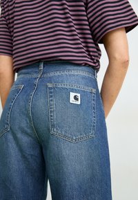 Jeans de denim azul con una cintura alta, que cuentan con un bolsillo trasero con una etiqueta blanca. La tela tiene una textura ligeramente desgastada. Camisa a rayas parcialmente visible.