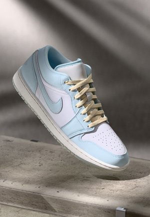 Lavtop sneaker med lyseblå og hvide paneler, beige snørebånd og hvid gummisål, placeret på en betonoverflade med en skyggefuld baggrund.