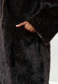 Faux fur frakke i mørkebrun med en tæt, blød tekstur. Har en sidelomme og brede, fyldige ærmer.