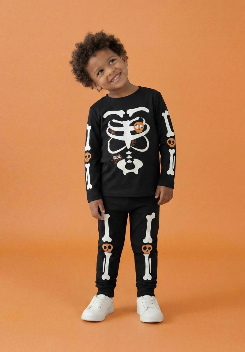 Jeune enfant souriant portant un costume de squelette noir avec des os blancs, des crânes et des motifs de toile d'araignée sur fond orange.