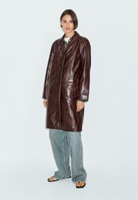 Trench in pelle marrone con colletto a punte, chiusura con bottoni e tasche laterali. Abbinato a jeans azzurri a taglio comodo e infradito.