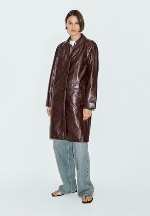 Trench in pelle marrone con colletto a punte, chiusura con bottoni e tasche laterali. Abbinato a jeans azzurri a taglio comodo e infradito.