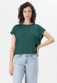 Žalias trumpa rankovė t-shirt su pasuktais rankovėmis ir laisvu siluetu. Lengva medžiaga su lygiu paviršiumi. Suporuota su šviesiai mėlynomis, aukštu juosmeniu džinsais.